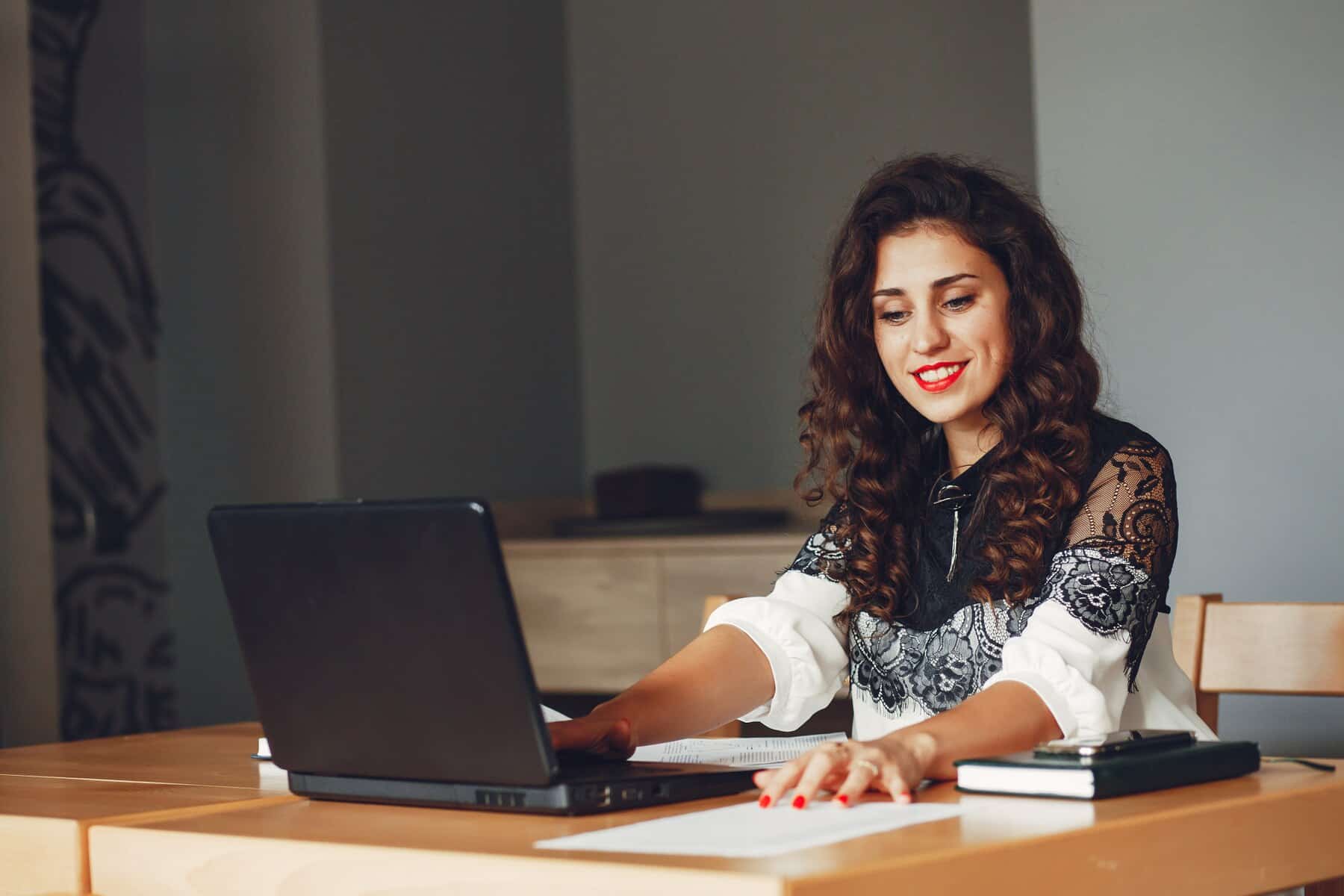 A woman with curly hair sits at a wooden desk, working on a laptop with papers and a notebook nearby.