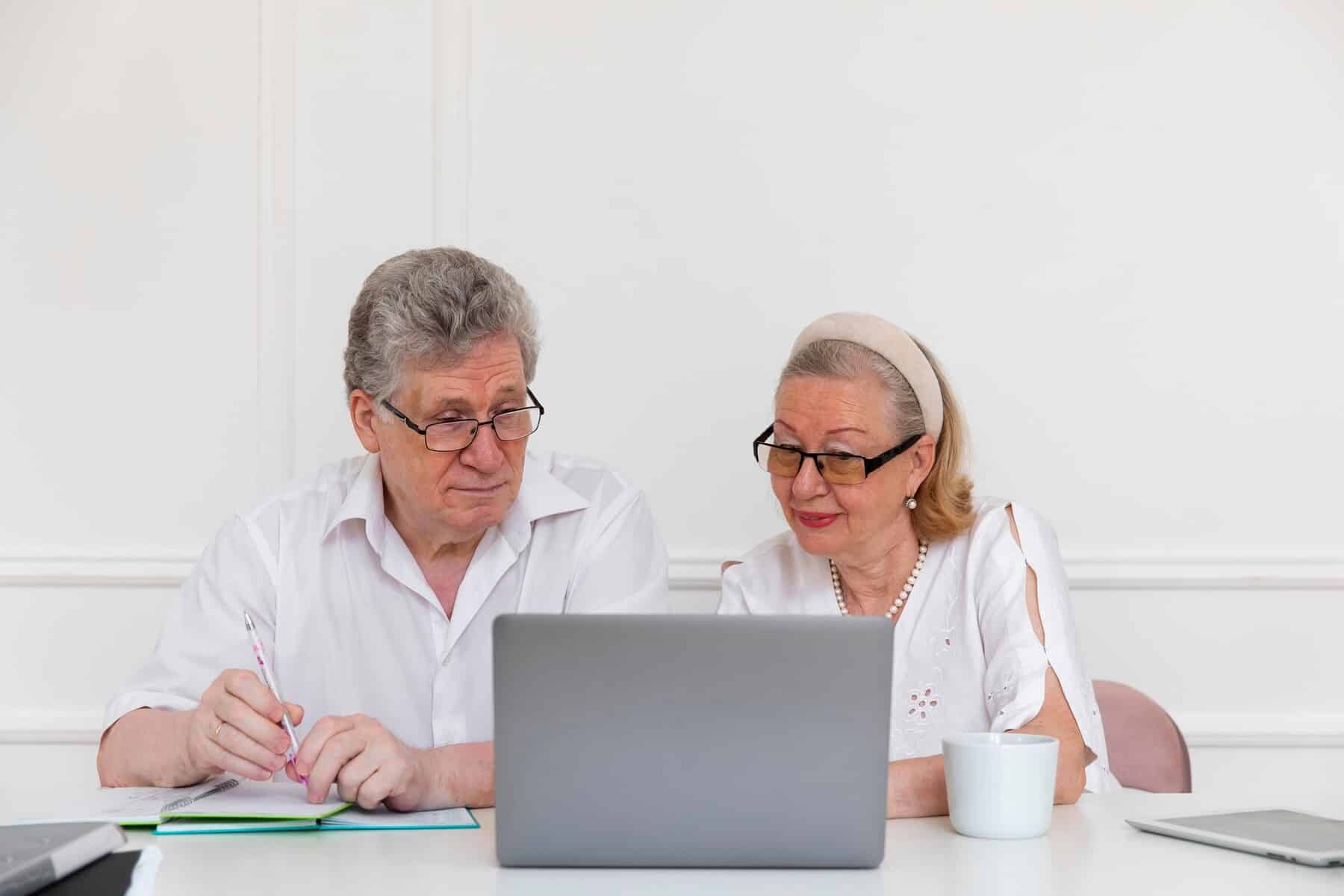 An elderly couple studies a laptop together at a table, with a notebook and a cup beside them in a bright, minimalistic room.