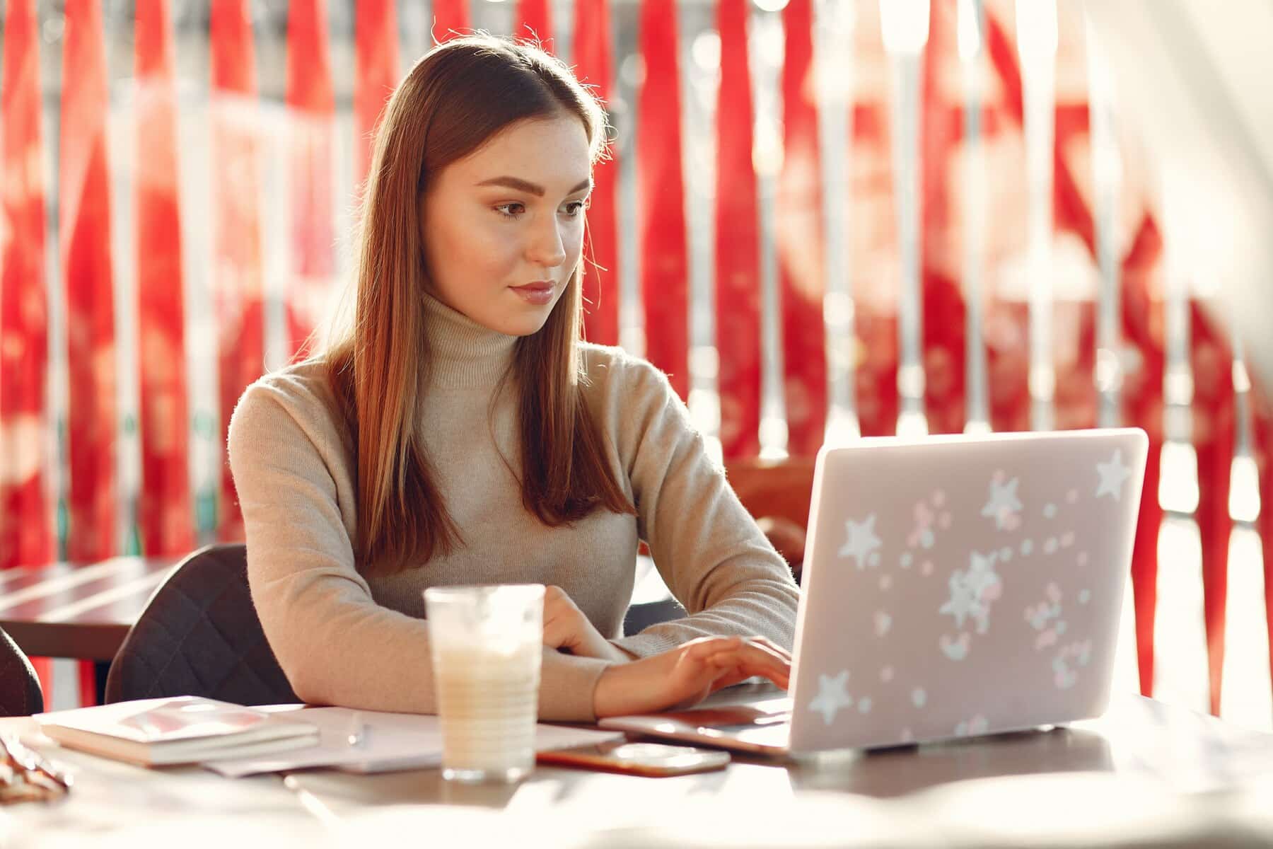 A woman sitting at a wooden table, focused on her laptop with a decorative cover, beside a glass of milkshake and notebooks.