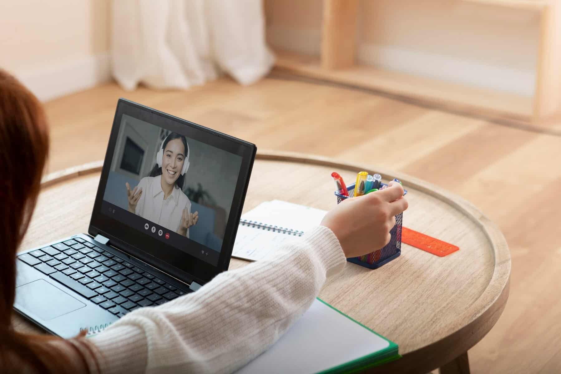 A person sits at a wooden table, engaging in a video call on a laptop, with a container of colorful pens beside them.