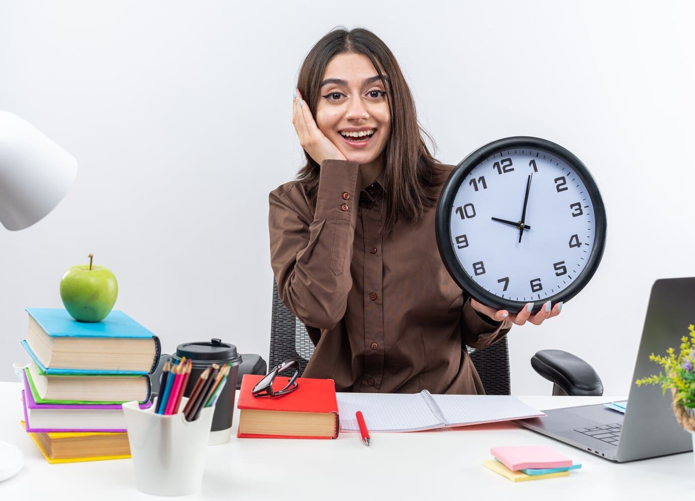 A young woman in a brown blouse smiling and holding a large wall clock at a desk surrounded by colorful books, a green apple, and stationery items, representing an organized work or study environment.