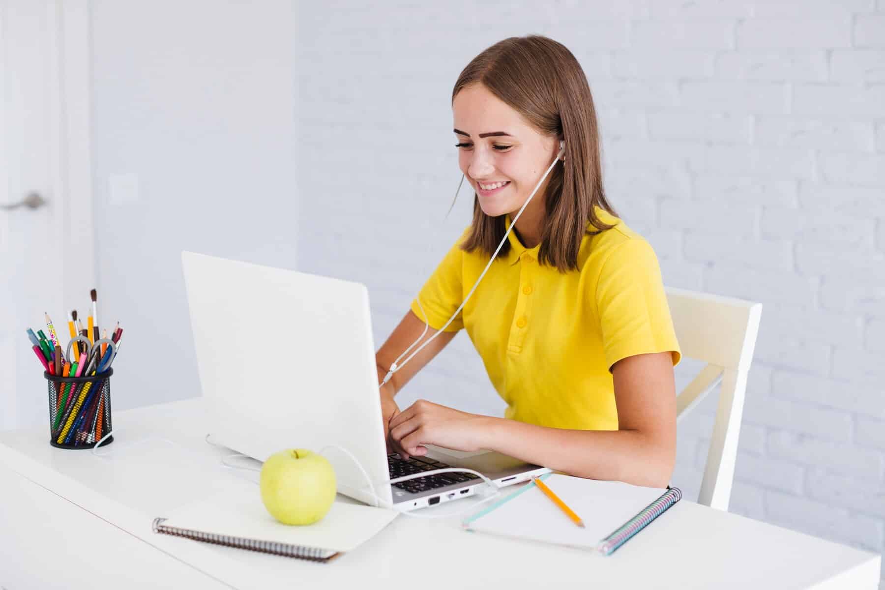 A person in a yellow shirt works on a laptop at a white desk, surrounded by colorful stationery and an apple.