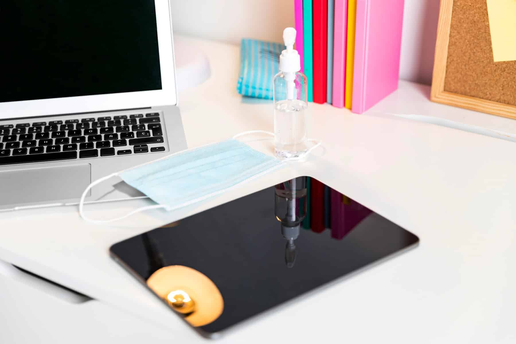 A modern workspace featuring a laptop, a surgical mask, hand sanitiser bottle, and colourful books on a white desk.