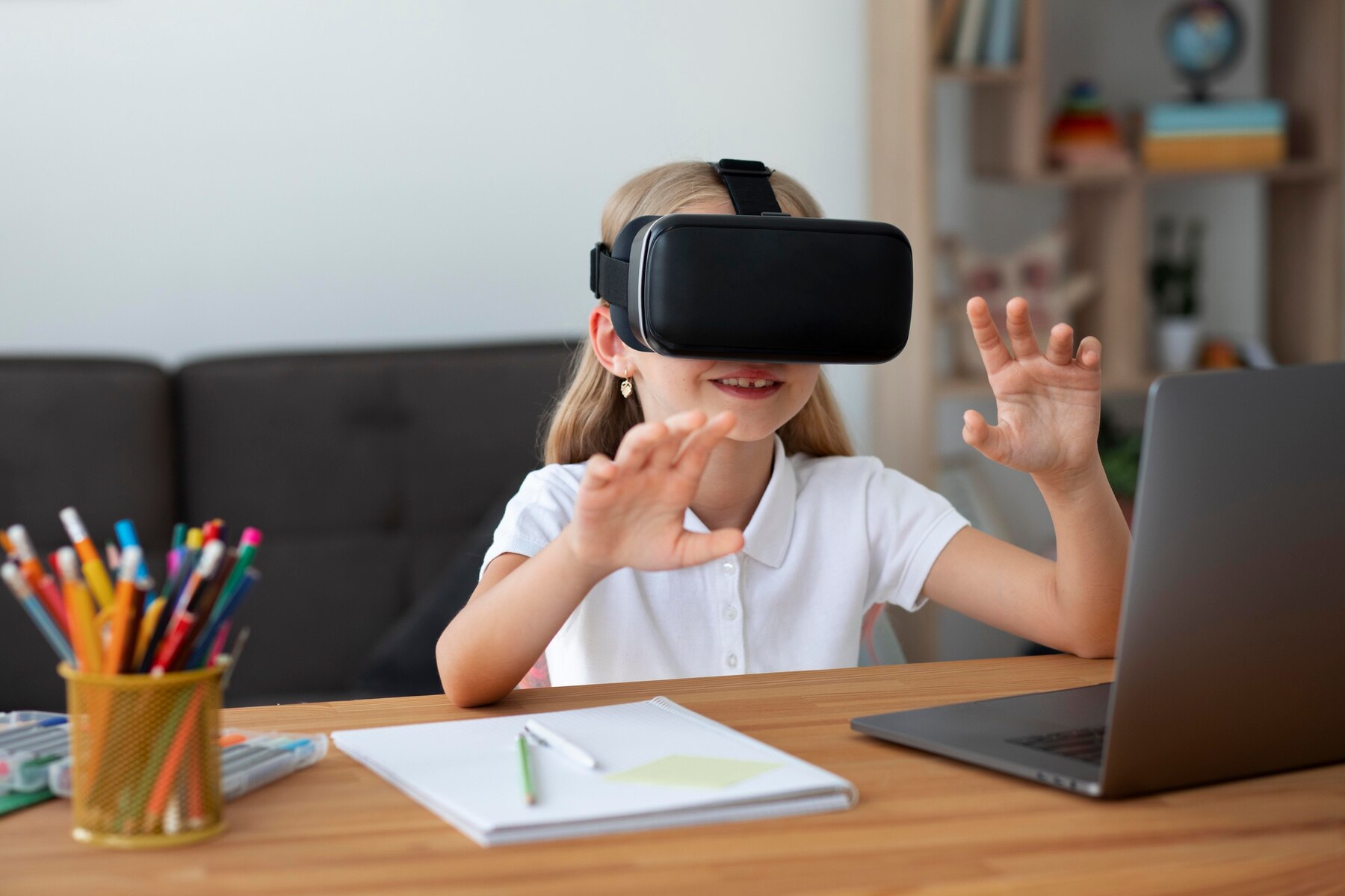 A young girl wearing a virtual reality headset smiles and gestures while seated at a desk with a laptop and art supplies.