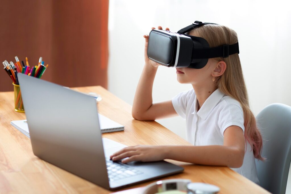 A child with long hair wears a VR headset while seated at a desk with a laptop, colorful pens, and a notebook nearby.