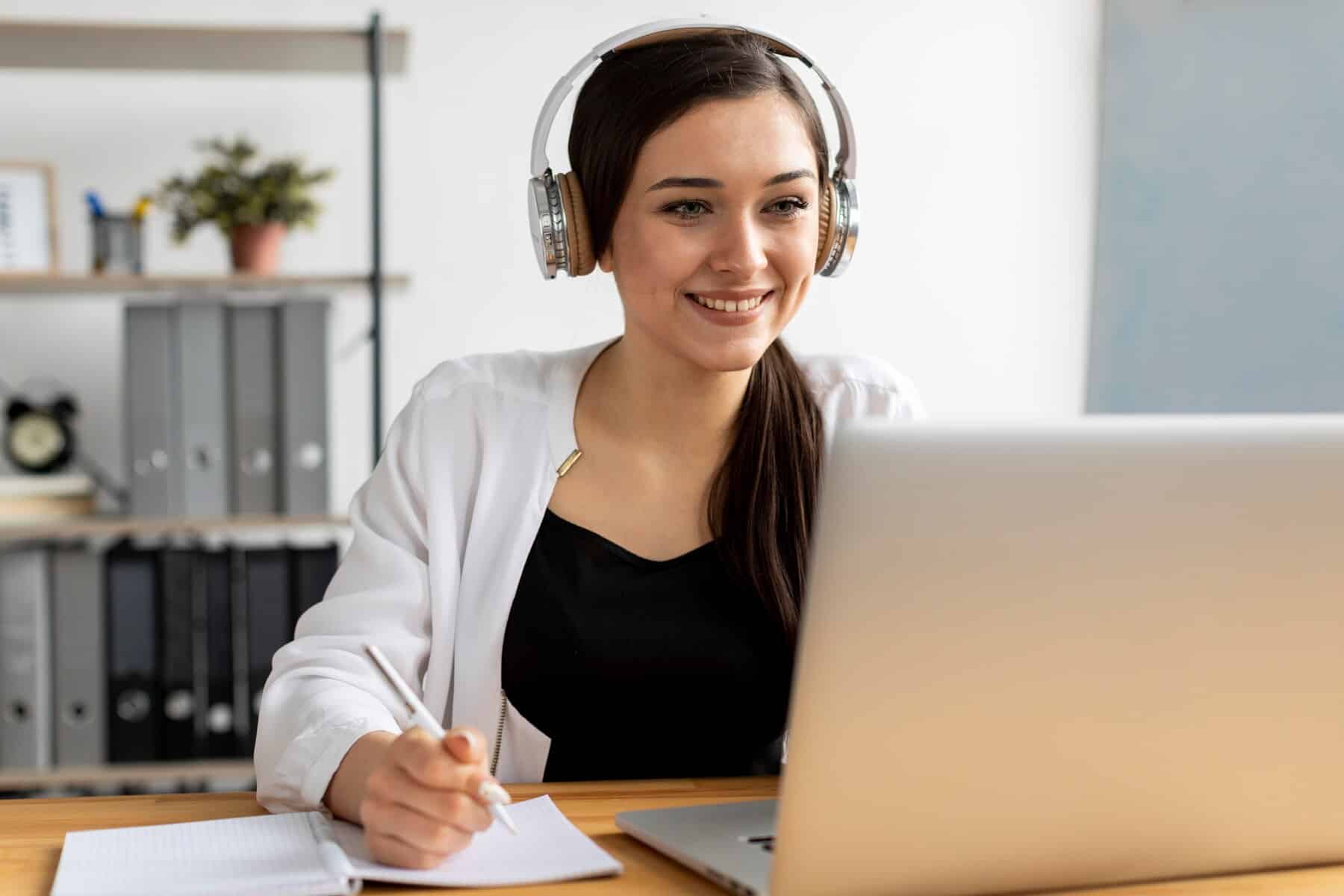A person wearing headphones sits at a desk, engaged with a laptop, taking notes in a notebook. Shelves with office supplies are visible behind.