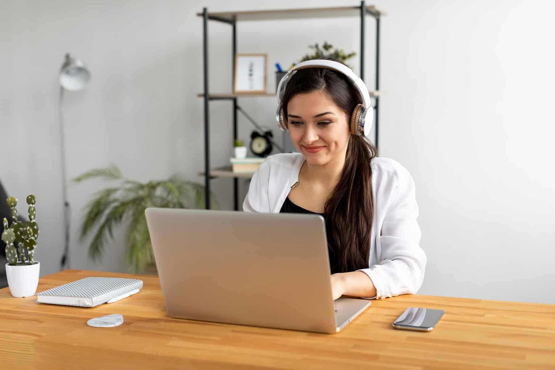 A woman wearing headphones works intently at a wooden desk with a laptop, notebook, and a small potted cactus in a bright room.