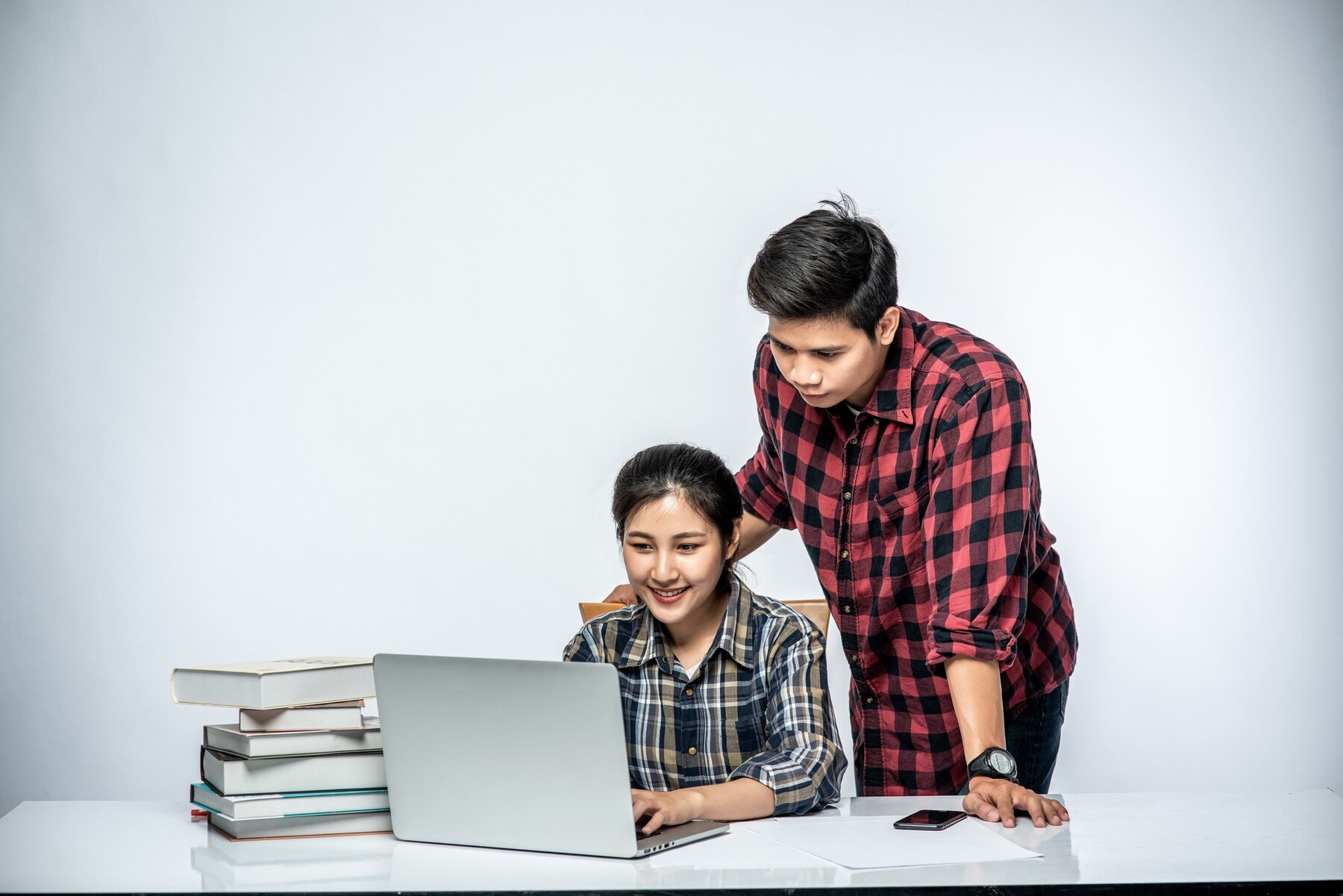 A student in a plaid shirt is focused on a laptop, while another person in a red checkered shirt offers guidance, surrounded by books.