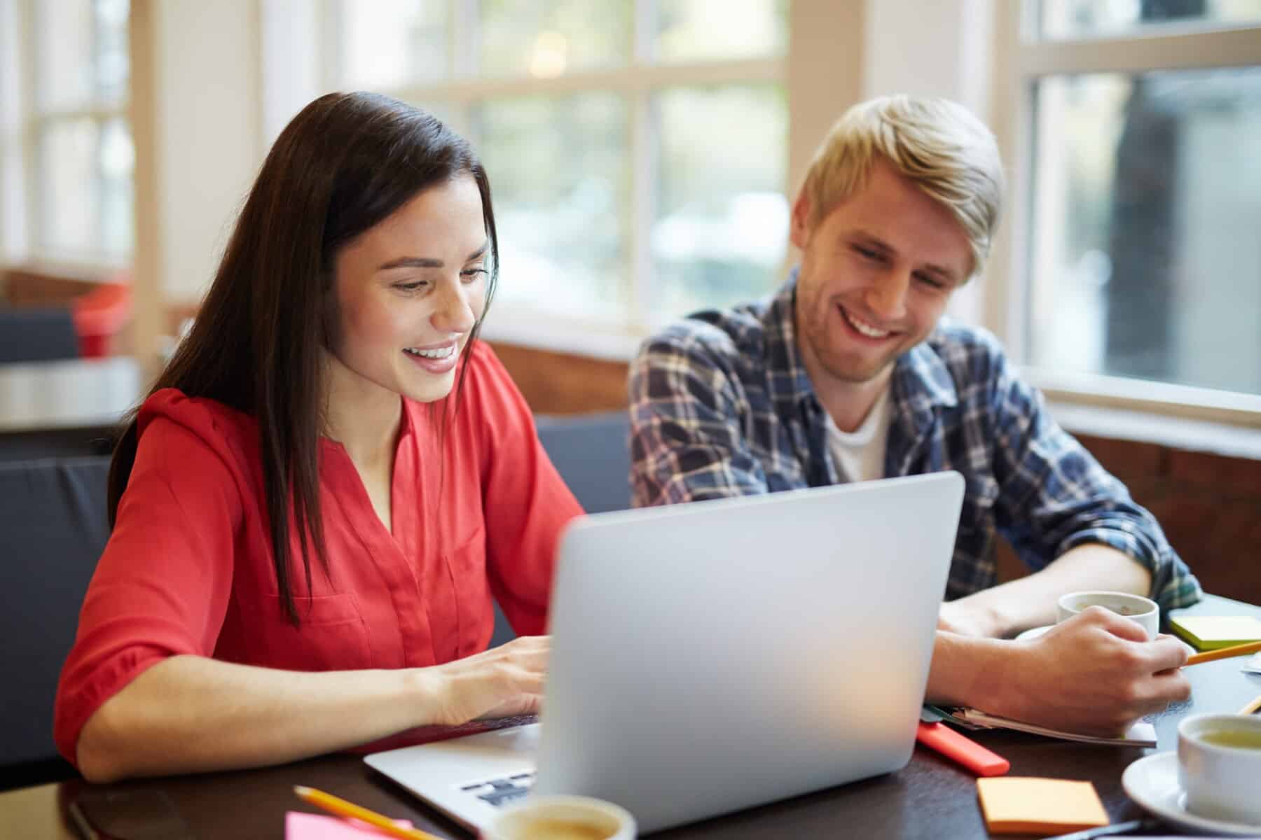 A woman in a red shirt works diligently on a laptop while a man in a checkered shirt sits beside her, both engaged in discussion.