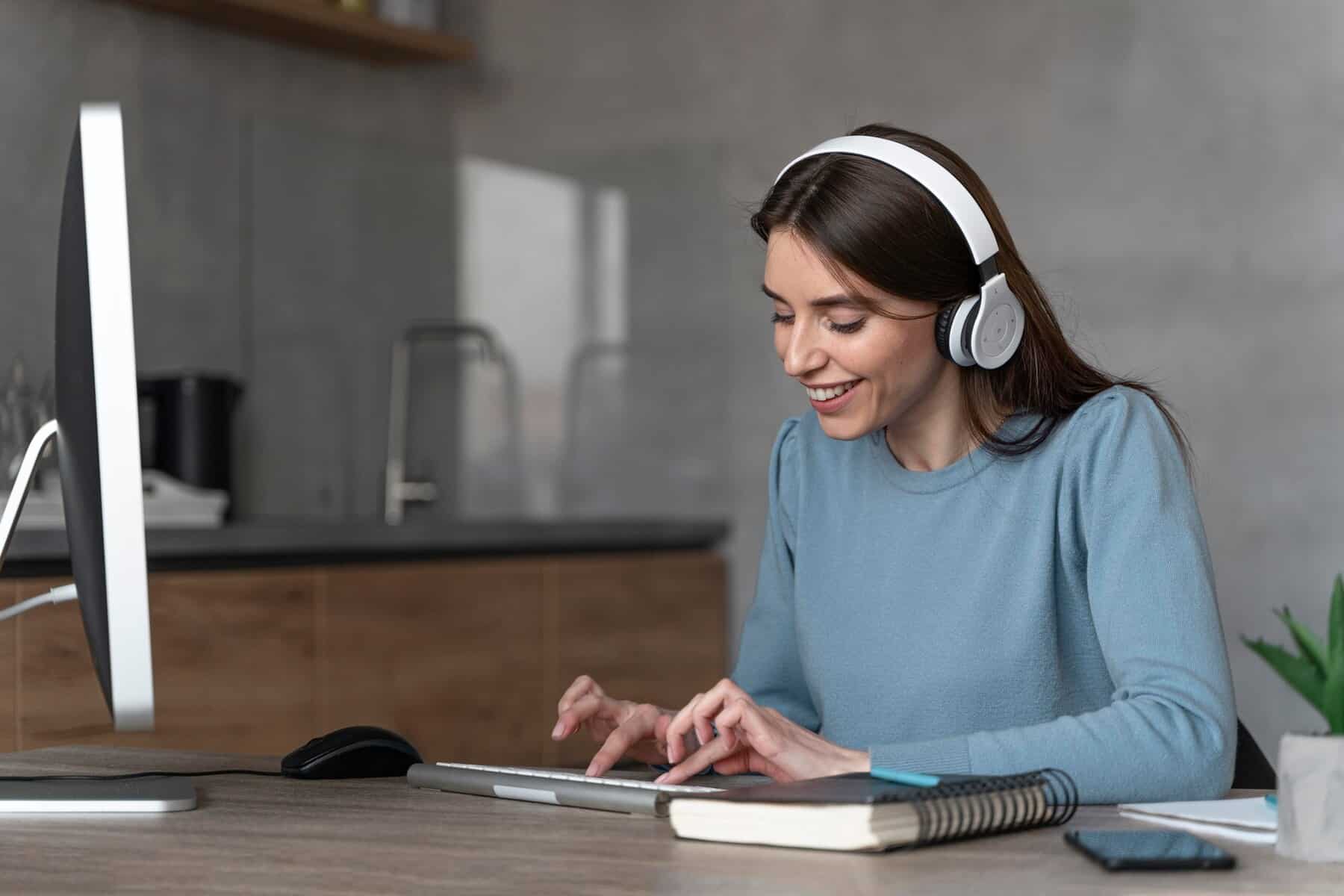 A person wearing headphones types on a tablet, sitting at a wooden desk with a computer and notebook, in a modern workspace.