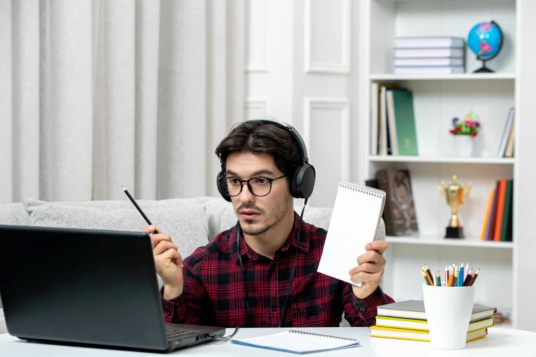 A student in headphones sits at a laptop, holding a notepad and pencil, surrounded by books and stationery in a cozy study space.