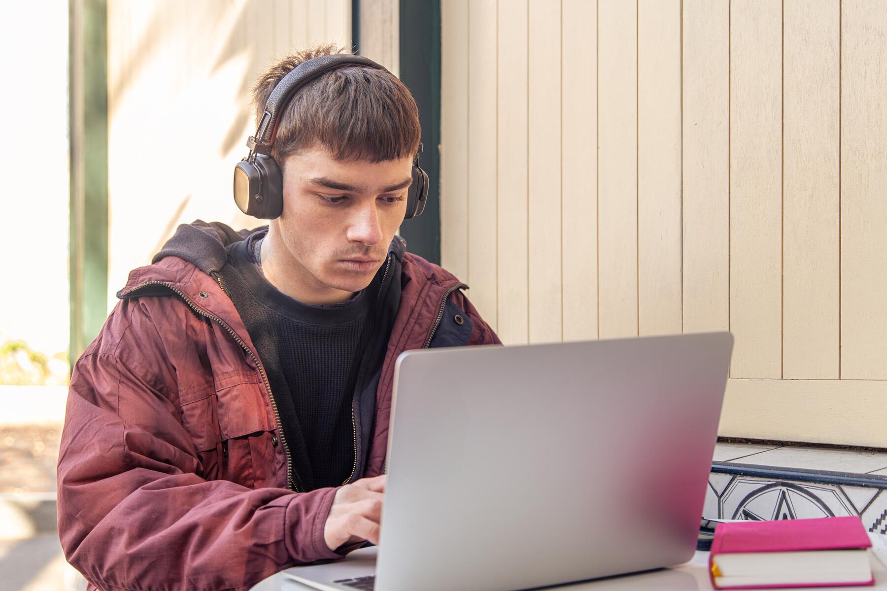 A person wearing headphones, sitting at a table, focused on a laptop, with a notebook and a pink notepad nearby.