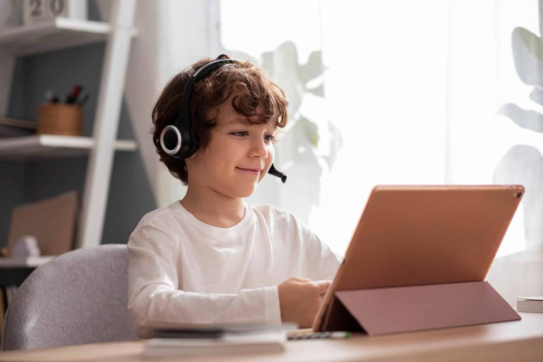 A child wearing headphones sits at a desk, focused on a tablet, with natural light streaming in through nearby windows.