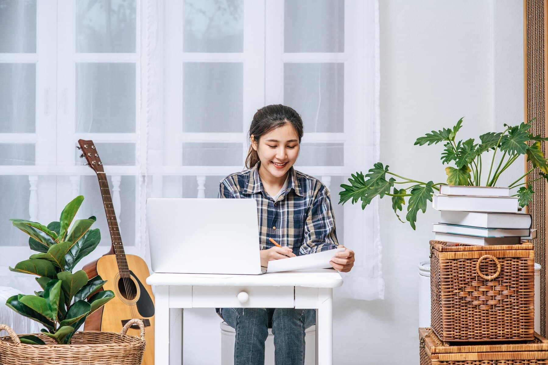 A person sits at a white desk with a laptop and notebook, surrounded by plants, a guitar, and books in a well-lit room.