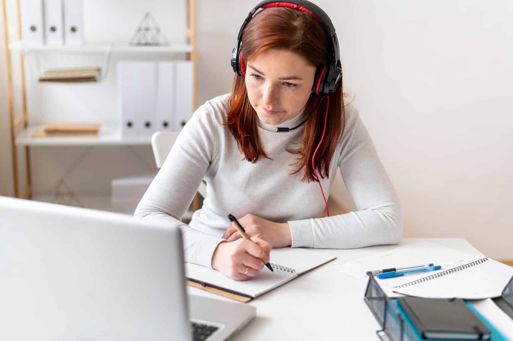 A person wearing headphones is focused on taking notes in a notebook, with a laptop and stationary on a desk.