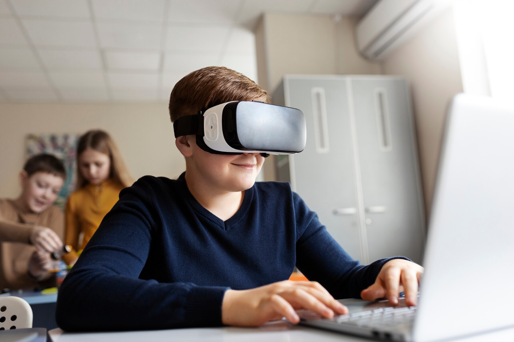 A student wearing a VR headset sits at a desk with a laptop, engaged in a digital experience, while peers are working nearby.