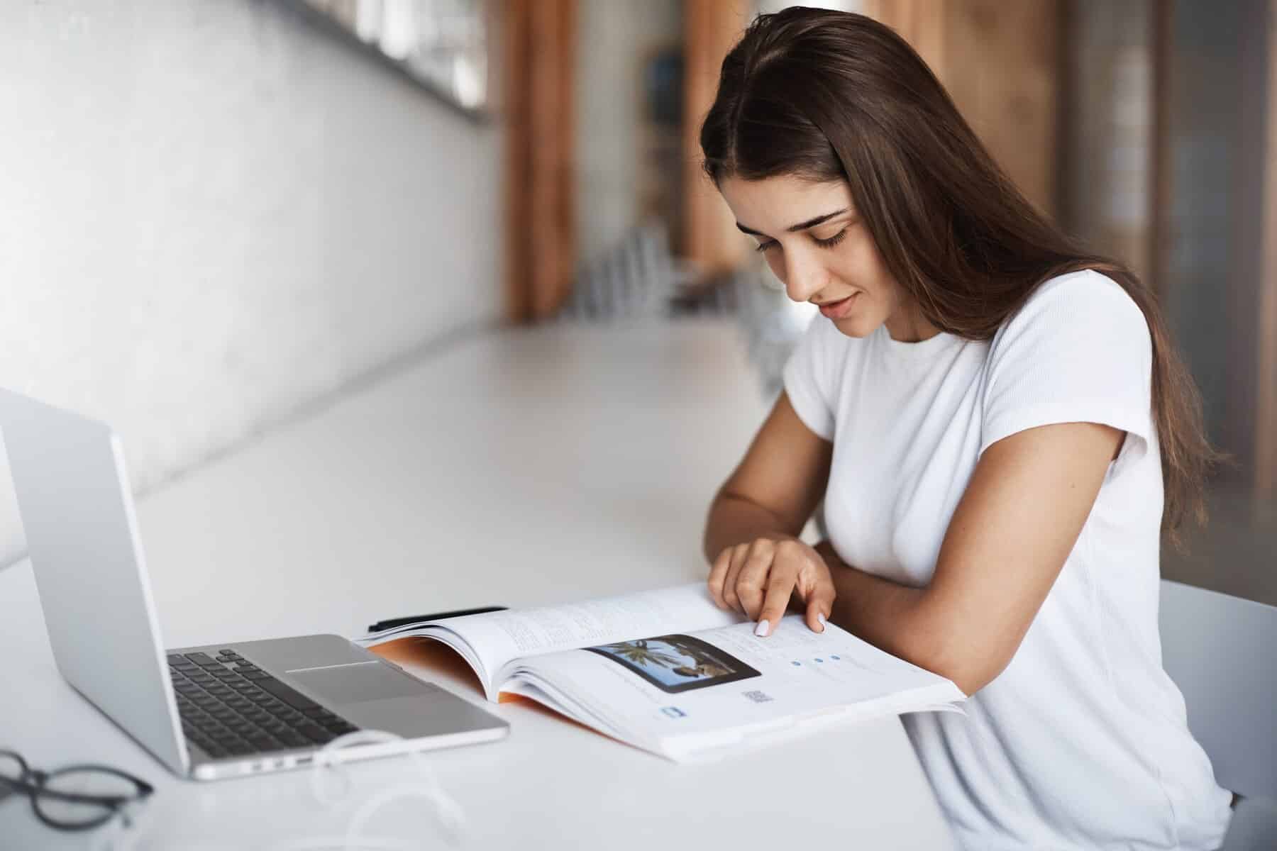 A person in a white t-shirt reviews a printed magazine on a desk, alongside an open laptop and eyeglasses. Bright, airy workspace setting.