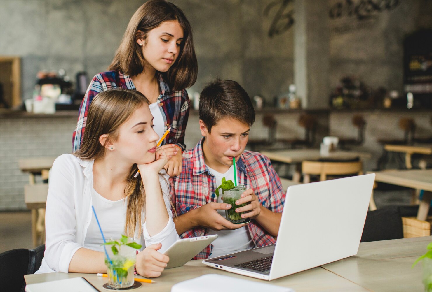 Three young people collaborate over a laptop in a cozy café, with drinks and a tablet on the table.