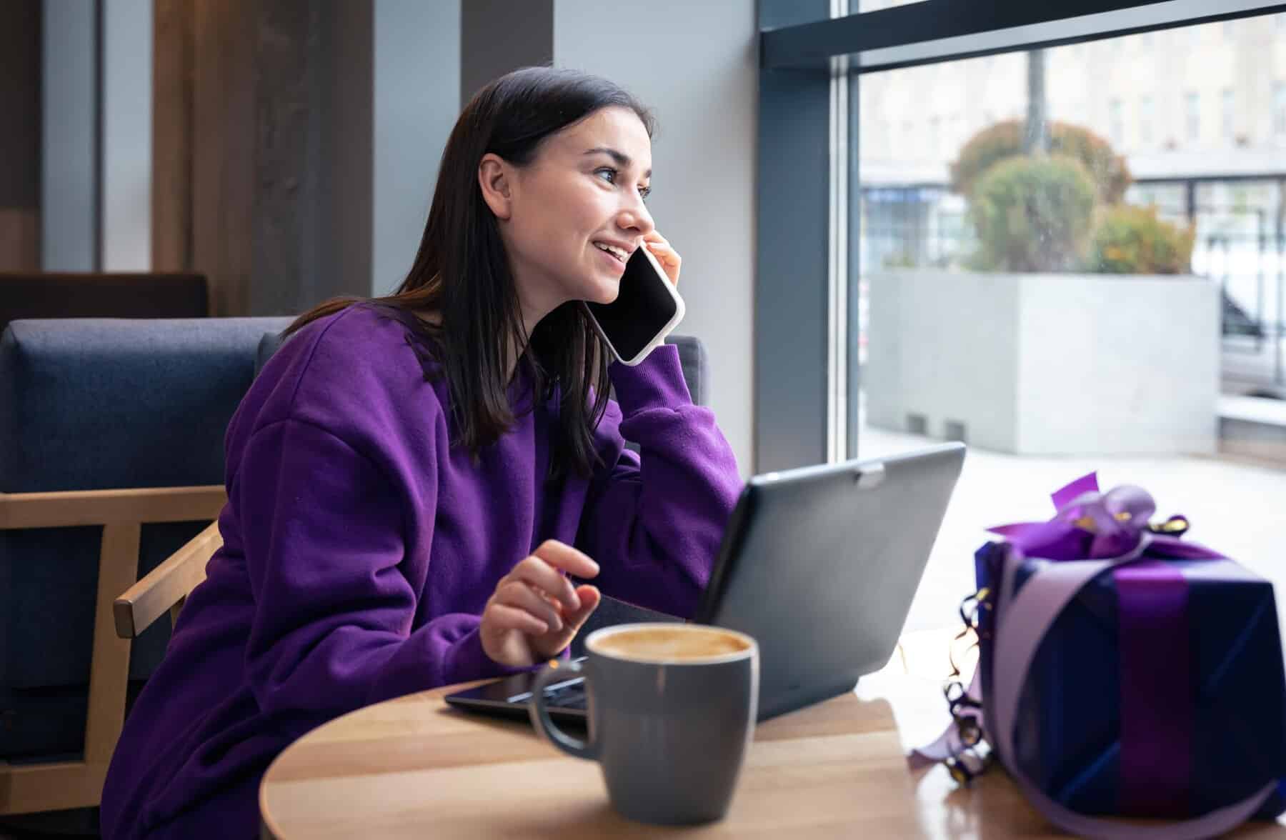 A person in a purple sweatshirt sits at a café table, talking on the phone, with a laptop and a coffee cup in front of them.