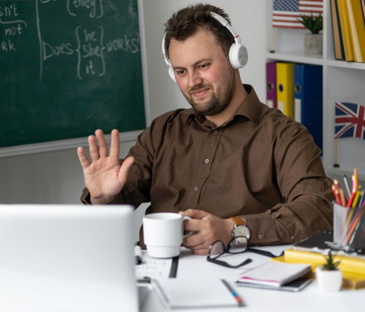 A man with headphones sits at a desk, waving and smiling, with a chalkboard and flags in the background.