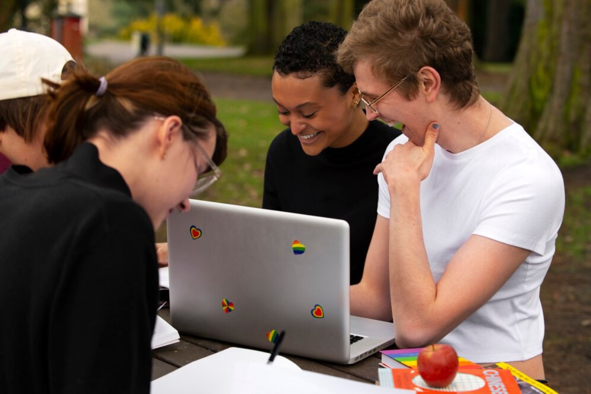 Three students smiling and looking at a laptop outdoors, with books and an apple on the table.