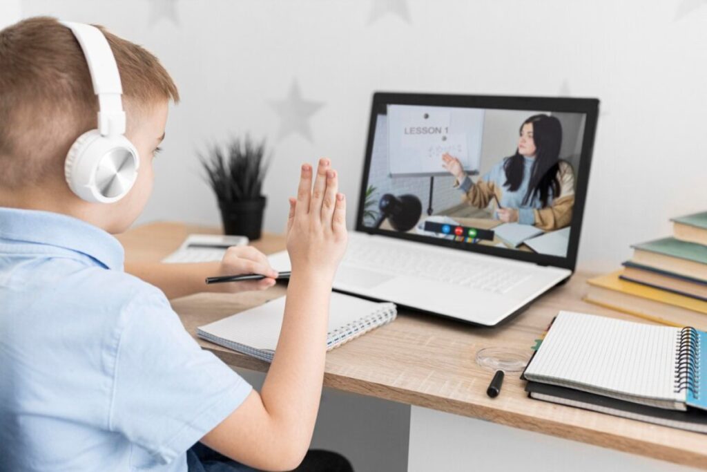 Young boy in headphones attending online class, raising hand to ask question.