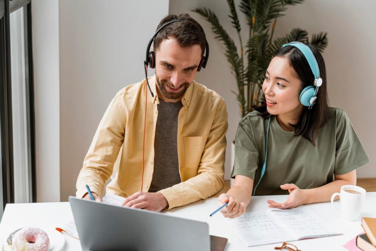 Two people with headphones collaborating at a desk with a laptop and notes.