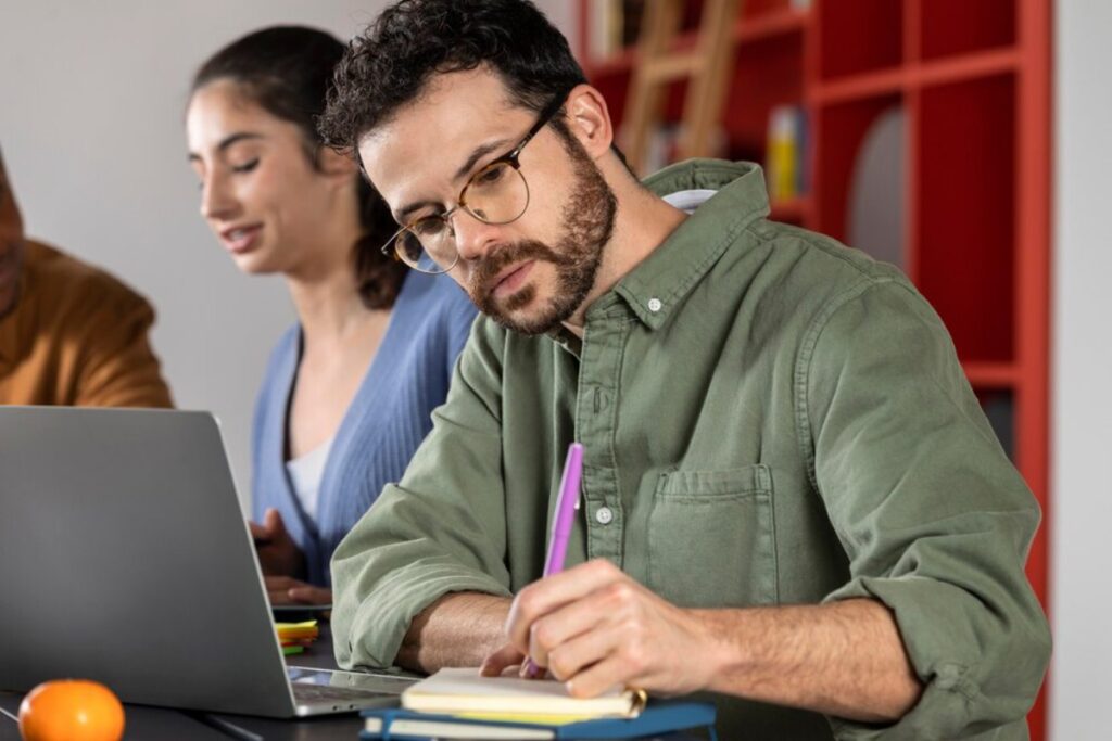 A man with glasses writing in a notebook, with two colleagues discussing in the background.