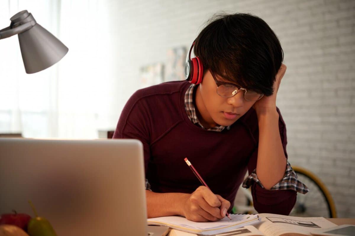Young man studying intently at a desk with headphones on, writing in a notebook, a lamp overhead.